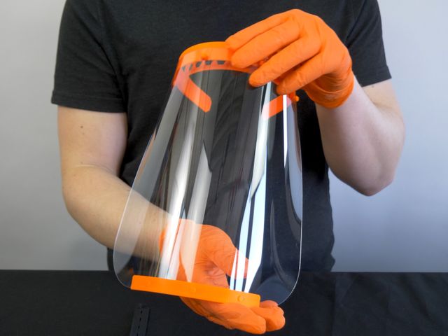 Hands in orange gloves holding a large clear face shield with stiff orange parts at the top and bottom